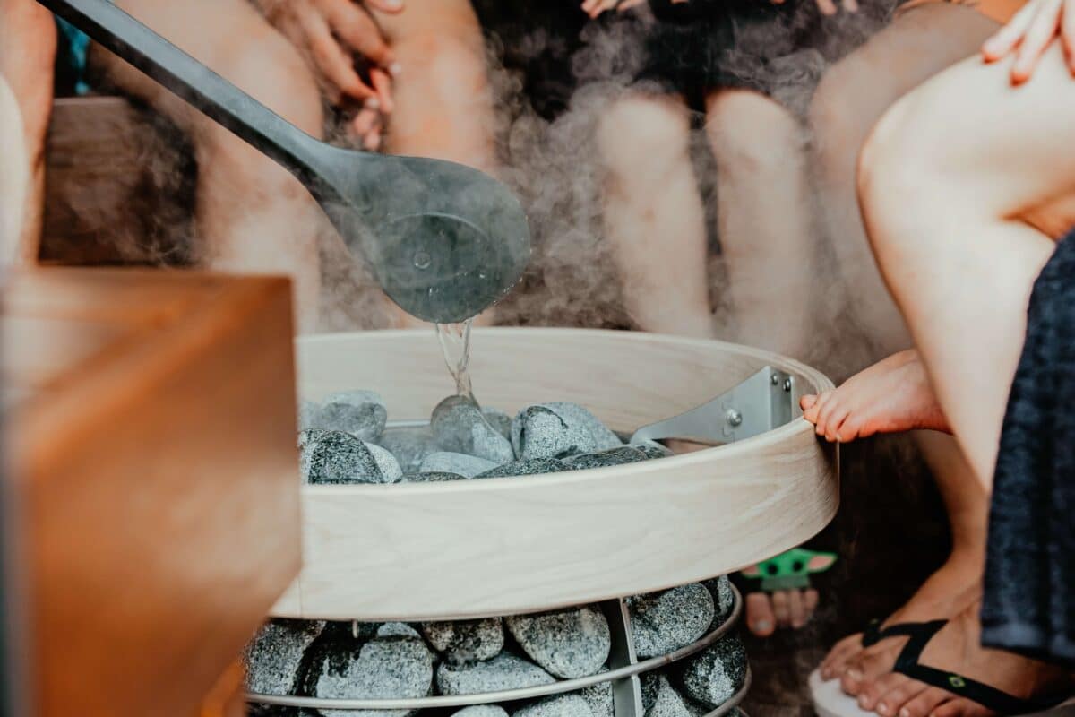 people's legs gathered around a sauna heater, with someone pouring water over the stones.